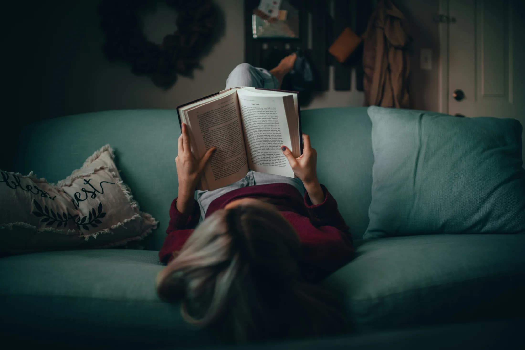 A person lying on a teal sofa reading a book
