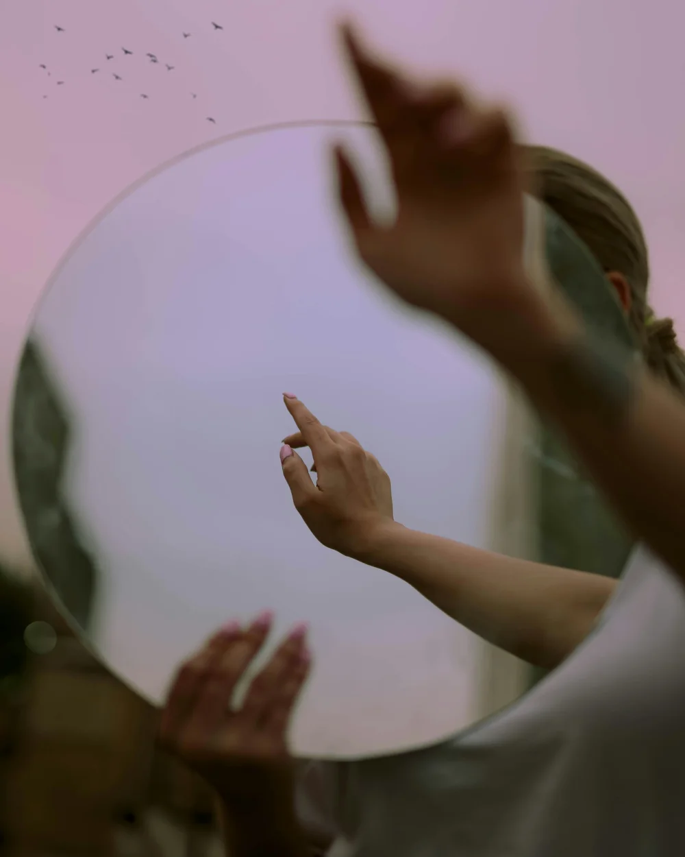 A hand reflected in a round mirror against a pink sky